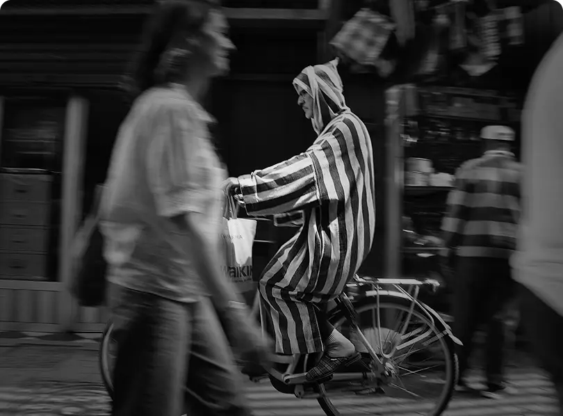 Person wearing a striped hooded garment rides a bicycle on a busy street with blurred pedestrians around.