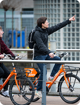 Two people on orange rental bikes, one pointing forward while stopped near a railing.